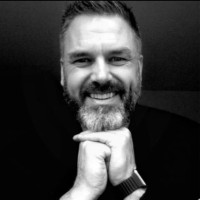 Black and white headshot of Gavin Neate, founder of welcoMe, smiling warmly with his chin resting on his clasped hands